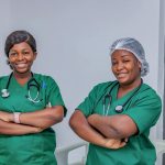 Two female nurses in green scrubs smiling confidently in a hospital room. Professional healthcare workers.